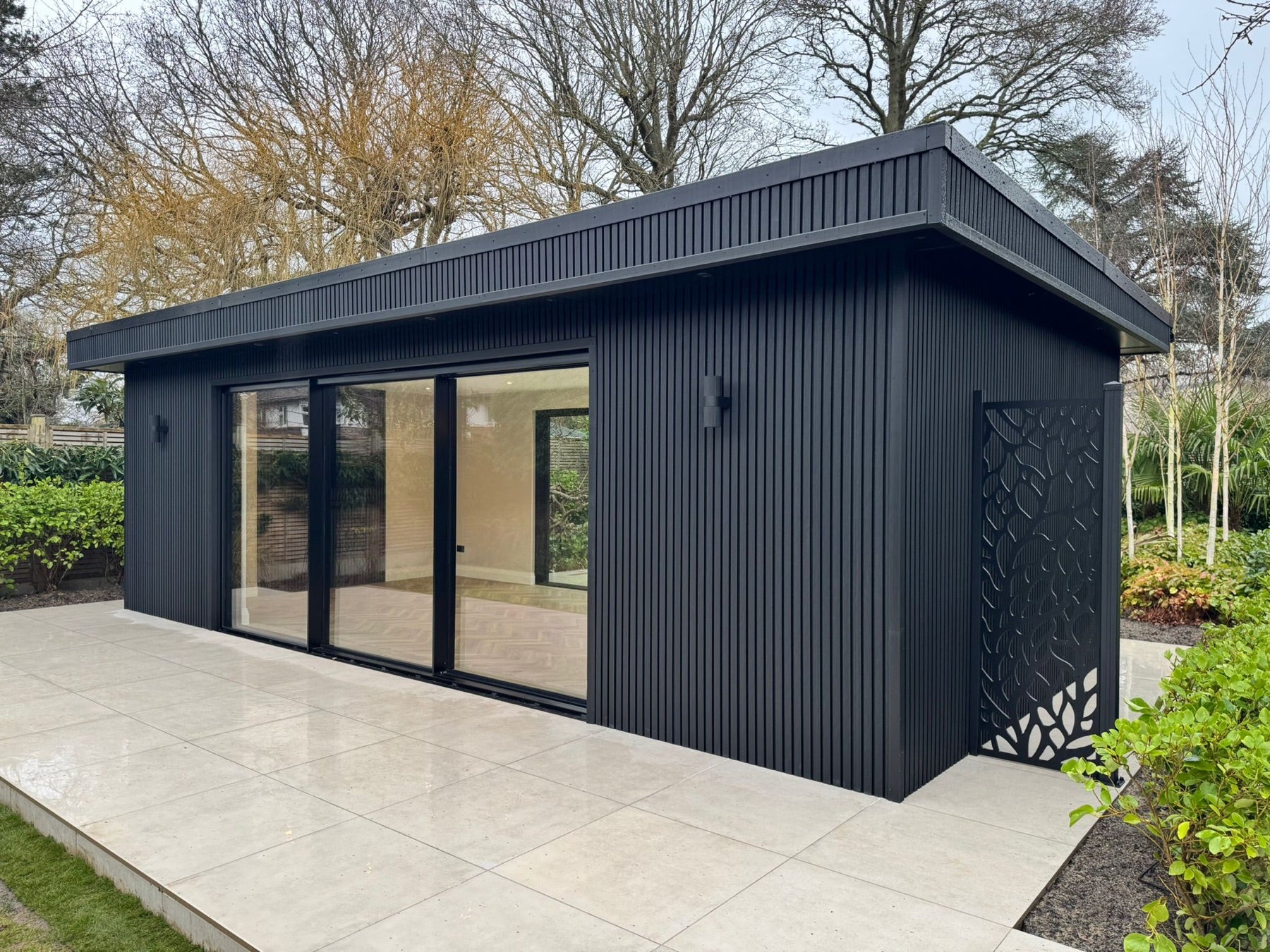 A garden room clad in black vertical slats, with sliding glass doors, wall lights and a decorative metal screen beside it, on a pale porcelain patio